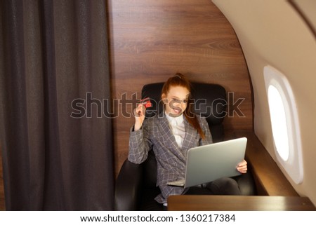 beautiful young girl with red hair is flying in a business class plane with a laptop and with a credit card in her hand
