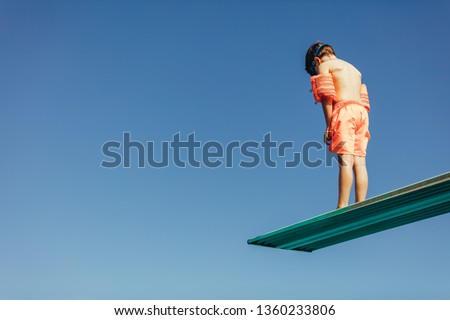 Low angle shot of boy with sleeves floats on diving board preparing for dive in the pool. Boy standing on diving spring board against sky. Royalty-Free Stock Photo #1360233806