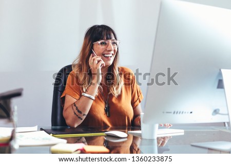 Smiling businesswoman talking on mobile phone while looking at a computer. Cheerful entrepreneur working in office sitting at her desk.