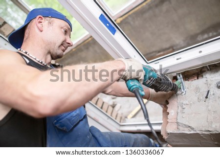 A male worker uses an electric perforator to drill a hole in a brick wall to install and fasten a PVC window. Royalty-Free Stock Photo #1360336067