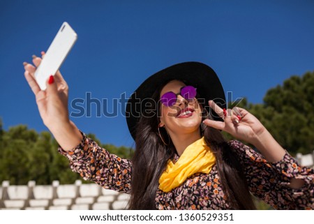 beautiful and young woman with hat, yellow handkerchief smiling and taking pictures of herself with her smart phone