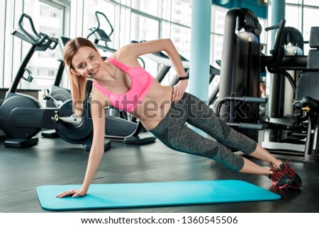 Young woman in gym healthy lifestyle standing on one arm on yoga mat in side plank looking camera smiling cheerful