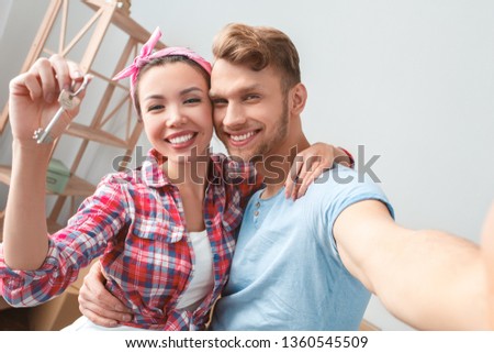 Young husband and wife moving to new place standing hugging taking selfie photos on smartphone looking camera smiling cheerful woman holding key from flat