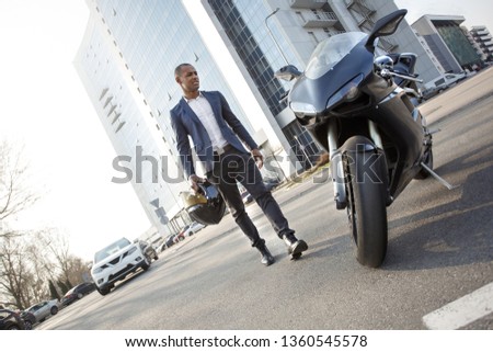 Young african american businessman coming to motorcycle carrying helmet squinting unsatisfied