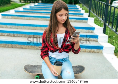 Girl with mobile phone brunette schoolgirl sitting in the summer in the city on the background of the stairs in a red shirt. Communication in the Internet, the application of social networks.