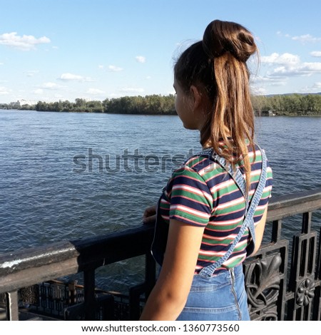 A young girl on the embankment of a large river looks at the water. Long hair gathered in a bun. Summer, striped short-sleeved clothing, jeans.