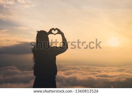 Silhouette of female holding hands in heart shape. Sunrise in the background. Summer vacation and travel concepts.