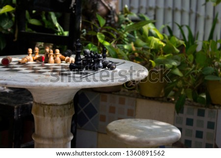 Outdoor stone chessboard with black and yellow figures. Competition and strategy concept. Defeat and fight concept. Chess wooden pieces on board in patio with green plants. Chess table in garden.