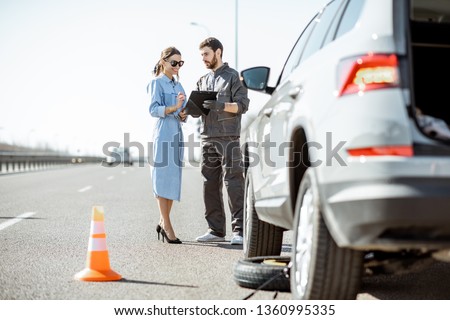 Road assistance worker signing some documents with woman near the broken car on the highway Royalty-Free Stock Photo #1360995335