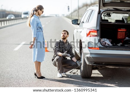 Road assistance worker helping young woman to change a car wheel on the highway Royalty-Free Stock Photo #1360995341