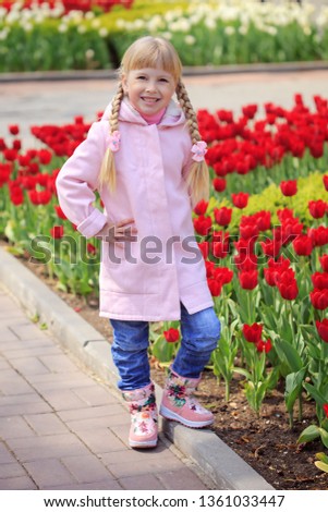 Cute beautiful little girl in a pink coat on a background of red and yellow tulips. Sunny day. Spring in the city