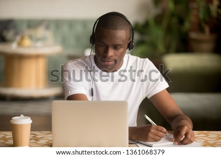 Focused african man wearing headphones watching webinar training making notes study online learning language on computer, black male student looking at laptop elearning in internet write information Royalty-Free Stock Photo #1361068379
