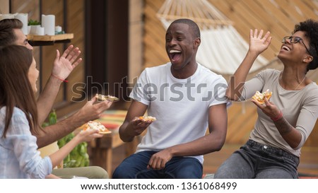 Happy diverse friends listening to young man telling funny story making laugh sharing dinner, multi-ethnic students people eat pizza in cafe terrace outdoor talking having fun together at meeting Royalty-Free Stock Photo #1361068499