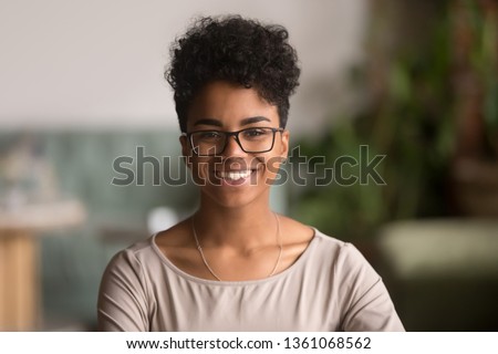 Head shot portrait of happy mixed race girl wearing glasses, smiling african american millennial woman posing indoor, pretty positive female student businesswoman young professional looking at camera Royalty-Free Stock Photo #1361068562