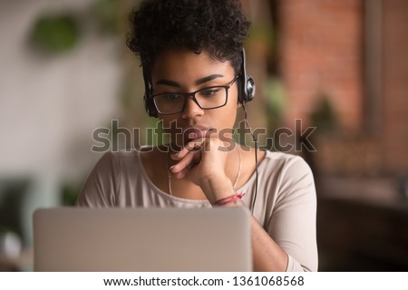 Focused african american girl student wearing headphones looking at laptop watching online webinar training course, serious mixed race woman learning english language on computer, internet e-learning Royalty-Free Stock Photo #1361068568