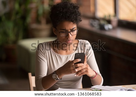 Upset confused african woman holding cellphone having problem with mobile phone, frustrated angry mixed race girl reading bad news in message looking at smartphone annoyed by spam or missed call Royalty-Free Stock Photo #1361068583