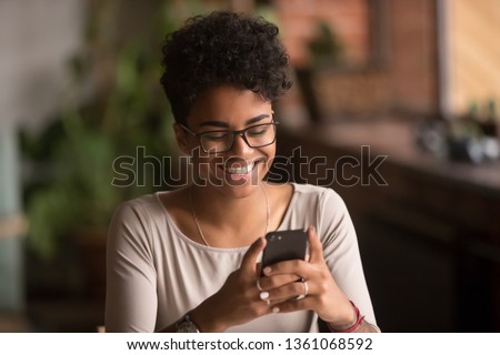 Happy millennial african american woman holding smartphone using ecommerce apps, playing mobile games, chatting in messengers or social media networks, surfing internet or texting message on phone