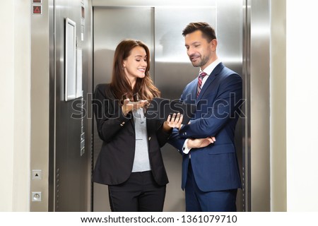 Smiling Young Businesswoman And Businessman Discussing While Using Digital Tablet Standing Near Elevator Royalty-Free Stock Photo #1361079710