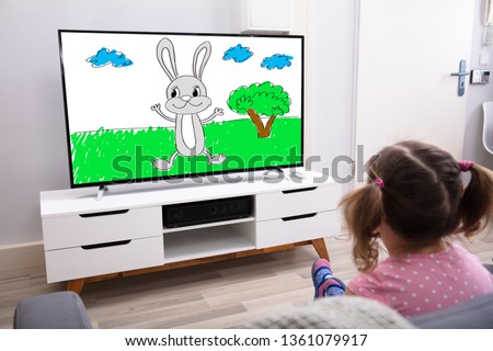 Rear View Of Innocent Girl Sitting On Sofa Watching Cartoon On Television
