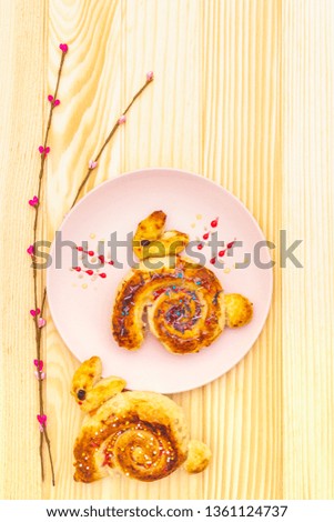 Fresh bun in the form of an Easter bunny. The concept of children's holiday bakery. In pink ceramic plate with artificial willow branches on wooden background, top view, close up