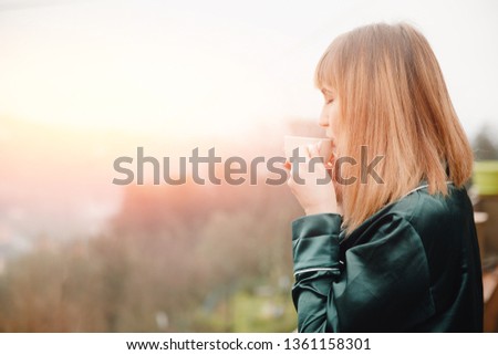 Beautiful young woman with cup coffee on terrace in green pajamas meets morning.