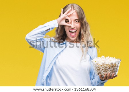 Beautiful young blonde woman eating popcorn over isolated background with happy face smiling doing ok sign with hand on eye looking through fingers
