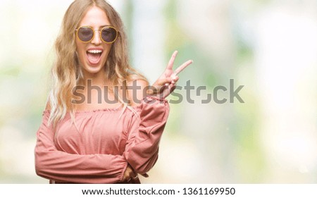Beautiful young blonde woman wearing retro sunglasses over isolated background smiling with happy face winking at the camera doing victory sign. Number two.