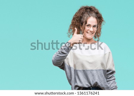 Beautiful brunette curly hair young girl wearing glasses over isolated background doing happy thumbs up gesture with hand. Approving expression looking at the camera with showing success.