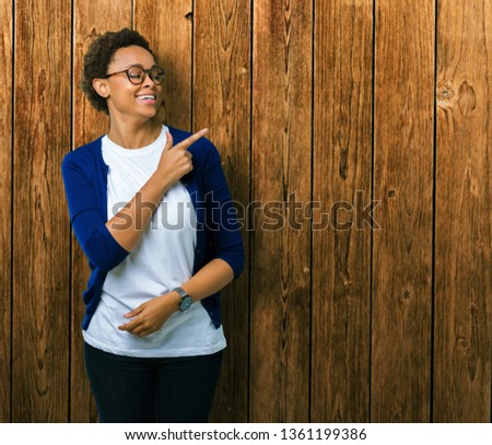 Young beautiful african american woman wearing glasses over isolated background cheerful with a smile of face pointing with hand and finger up to the side with happy and natural expression on face