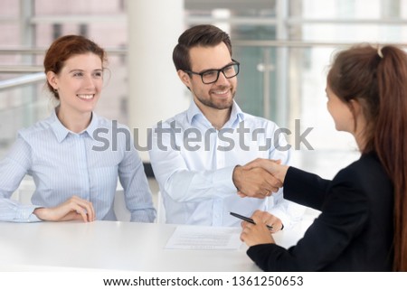 Happy young couple signing mortgage insurance investment contract handshake broker realtor insurer, smiling services buyers shake hand of bank agent saleswoman making financial deal at meeting