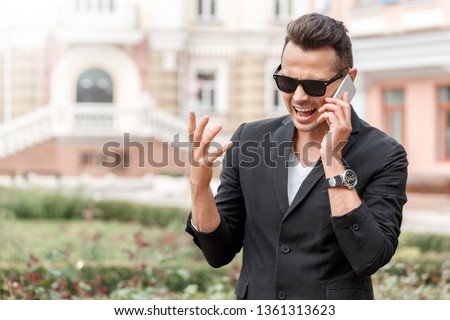 Young stylish man wearing suit and sunglasses outdoors standing on the city street shouting angry at business partner on smartphone clsoe-up