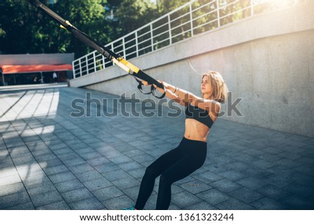 Young beautiful blonde woman using suspension straps in her exercise routine outdoors in the evening