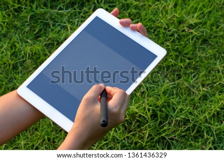 Hand holding tablet and using stylus on green grass background with soft sun light.