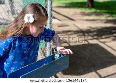 Little Girl in Blue Dress Using Drinking Fountain at Park by Baseball Diamond