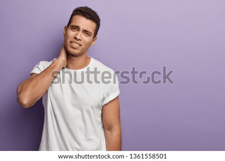 Studio shot of tired overworked young man needs massage, feels pain in neck, sitting hardworking long time, wears t shirt, expresses negative emotions, isolated over purple wall, free space.