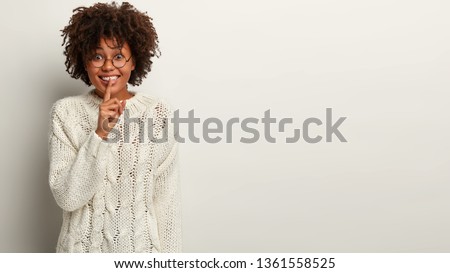 Cheerful dark skinned female shows hush sign, keeps fore finger over lips, smiles happily, tells pleasant secret to close friend, wears white winter outfit, stands in studio, blank space for your text