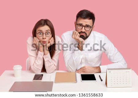 Photo of displeased office worker and his female partner look surprisingly and sadly at camera, tired of working, use modern gasgets, pose at workplace, drink takeaway coffee, isolated over pink wall