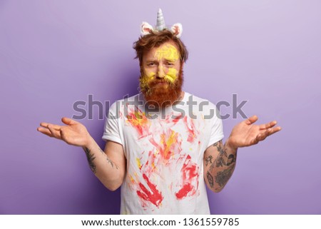 Unaware bearded young man dirty with colourful watercolours, spreads hands with indecisive expression, wears unicorn horn and ears, has white t shirt with spots, isolated over purple background Royalty-Free Stock Photo #1361559785