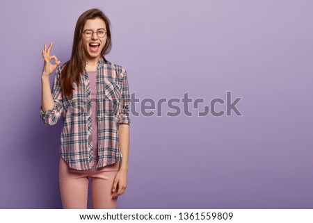 Playful female makes okay sign, feels happy after making deal, blinks eye, expresses positive emotions, dressed in casual checkered shirt, isolated over purple wall, blank space. Body language