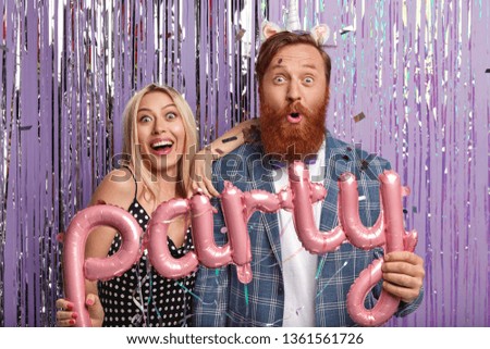 Fun party girlfriend and boyriend smile gladfully, hold balloon letters, stand closely, express surprise as observe excting performance, stand over decorated studio wall. Awesome celebration Royalty-Free Stock Photo #1361561726