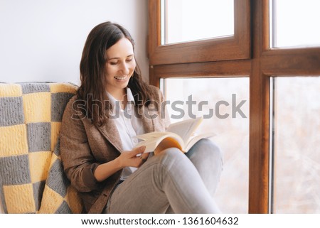 Photo of relaxed woman sitting on a couch near a window with a book. girl reading at home