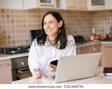 Photo of happy young woman with laptop, credit card and smartphone looking forward in kitchen at home