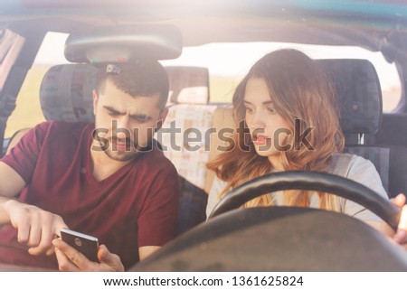 Puzzled couple sits in automobile, bearded man holds smart phone, uses online maps, tries to find way, being lost, have journey on weekend. Family in automobile chek email while parking on road side.