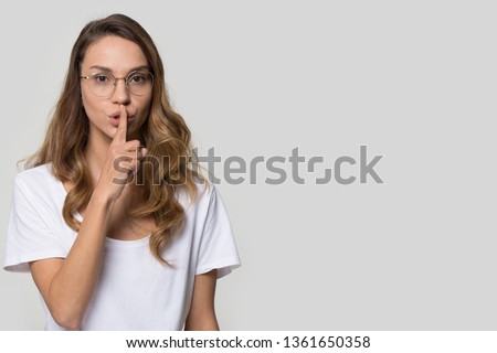 Attractive woman in glasses makes hush gesture silence symbol finger on lips looking at camera posing over grey wall background studio shot, copy space for your advertisement text, concept of secret