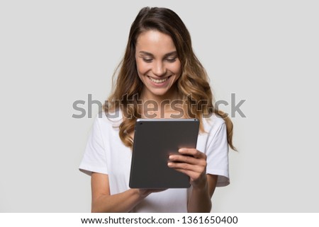 Smiling woman in white t-shirt over gray background holding wireless gadget looking at digital tablet screen having fun surfing internet chatting online with friends reading media news, concept image