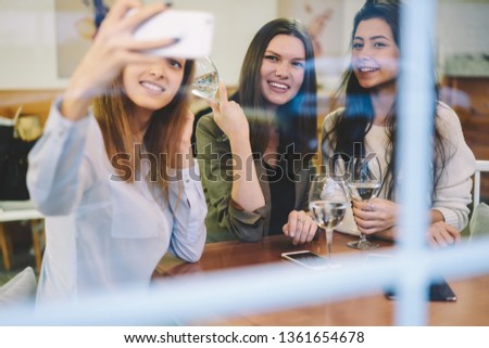 Young smiling hipster girl holding modern smartphone gadget in hand and clicking selfie photo for posting at own web blog during leisure time with female friends for drinking wine and communicate