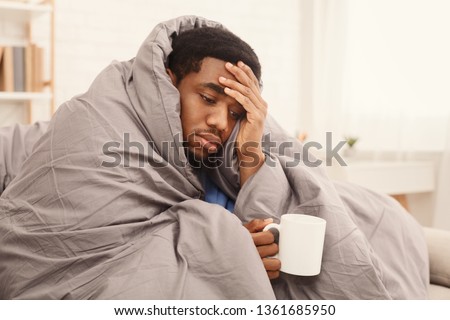 Folk medicine. Sick african american guy with fever wrapped in warm blanket drinking hot tea, free space Royalty-Free Stock Photo #1361685950