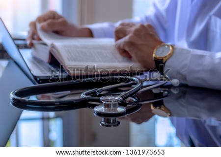 Male doctor in white lab coat concentrate on reading book, with  laptop computer and stethoscope on the desk at hospital.
