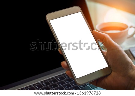 Mock up image of business person hand holding mobile smartphone with empty white screen and working on laptop computer with cup of coffee on table. 