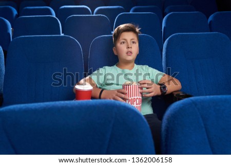 Teenager sitting in movie theatre in comfortable dark blue chair. Handsome boy holding popcorn bucket, red paper cup with fizzy drink. Concept of entertainment and leisure.